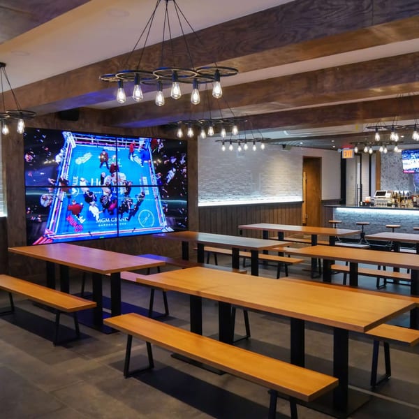 Interior of Boardwalk Beer Garden with long wooden tables, benches, and a large screen showing a boxing match.