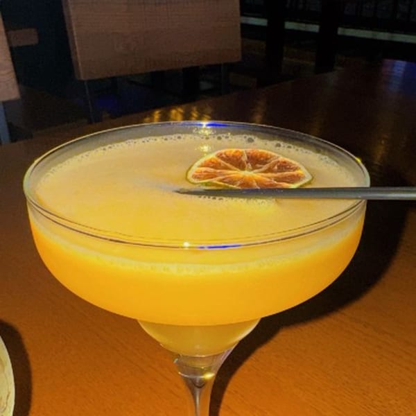A bright orange cocktail in a martini glass, garnished with a dried orange slice and a straw.