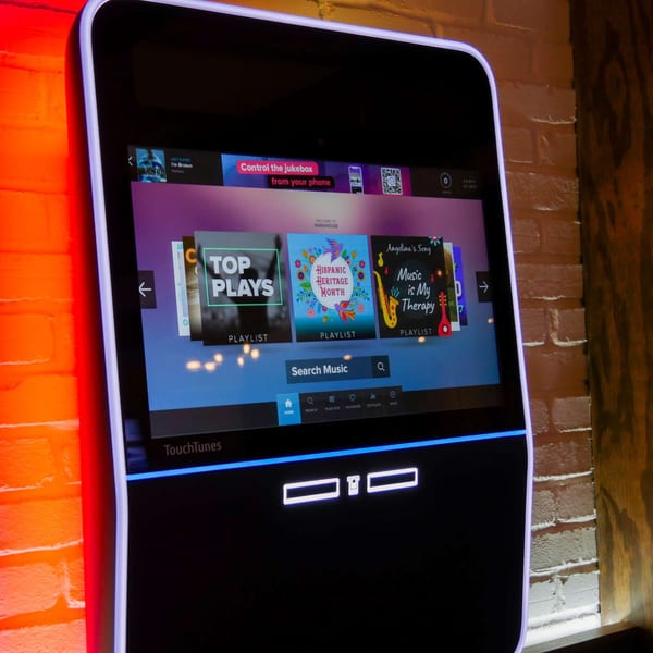 A TouchTunes digital jukebox displaying various music playlists, illuminated by red and blue lights.