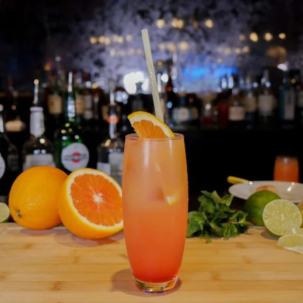 A vibrant orange cocktail with an orange slice and straw, surrounded by fresh citrus and mint.