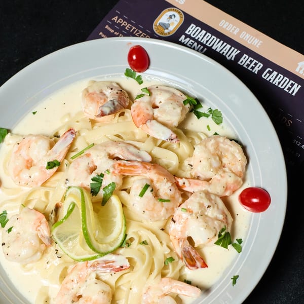 Creamy shrimp fettuccine pasta with lime, parsley, and cherry tomatoes, next to a Boardwalk Beer Garden menu.