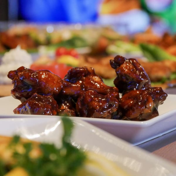 Sticky, glazed chicken wings on a white plate, with other appetizers blurred in the background.